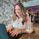 Terra Brown of Adventures by Terra smiles while typing on a laptop at a stylish indoor venue during Summer Camp at Sea. She wears a sleeveless white top and sits beside a frosty iced coffee in a stemmed glass, with a decorative mirrored wall in the background.