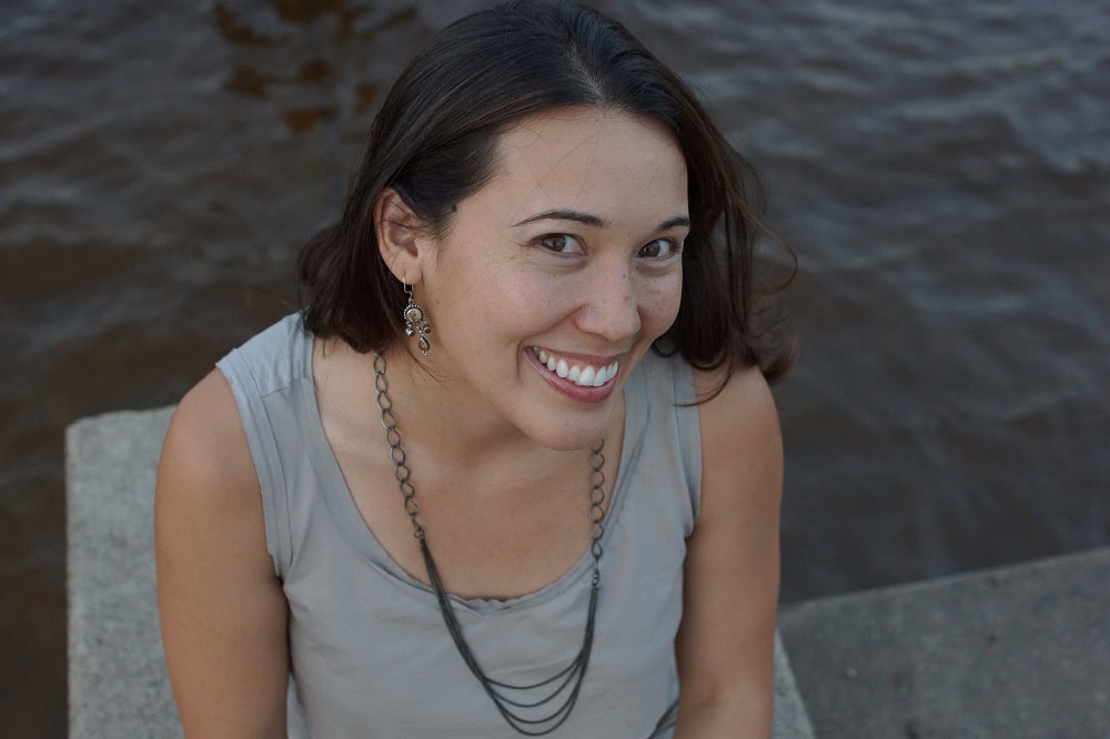 Steph Lee, founder of Host Agency Reviews, smiles at the camera while sitting near the edge of a body of water. She wears a sleeveless gray top, long chain necklace, and dangling earrings, with soft waves visible in the background.