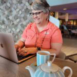 Carri Arman of Timeless Stones Travel sits at a table onboard a cruise ship smiling while working on a laptop, with a teapot and cup in the foreground, capturing a productive moment during a travel agent content retreat at sea.