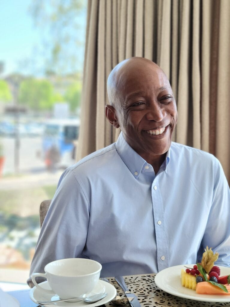 Smiling man in a light blue button down shirt seated at a breakfast table with fruit and coffee in front of him. Warm and approachable brand photoshoot image for travel and lifestyle business content.