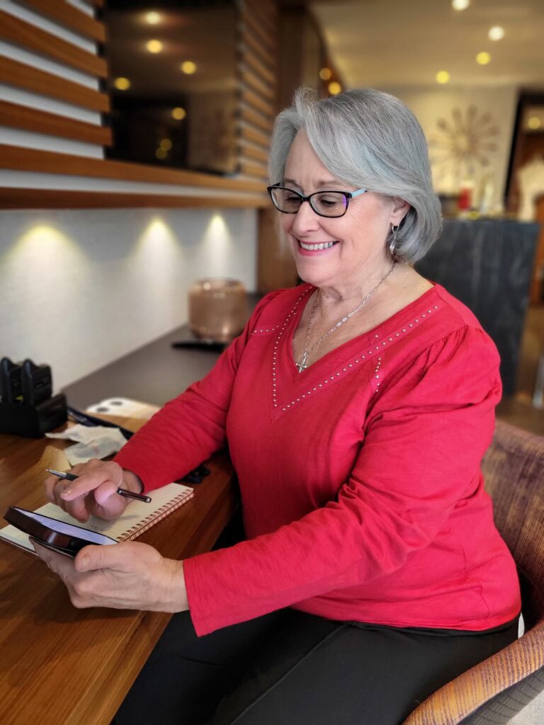 Woman in a bright red top smiling while using a smartphone and holding a pen beside an open notebook at a wooden desk. Professional travel agent content shoot image showing planning and communication.