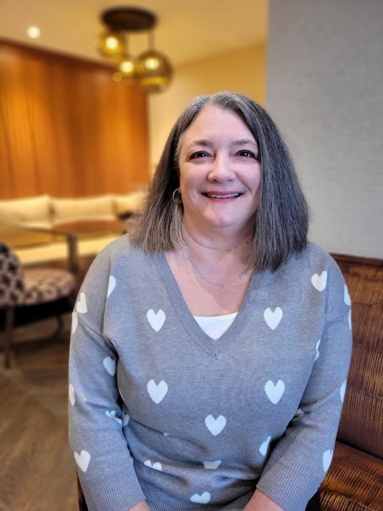 Woman in a gray sweater with white heart patterns smiling while seated on a couch in a softly lit lounge. Relaxed business photography portrait captured during a travel agent content shoot.