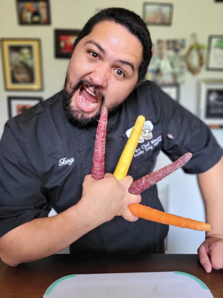 Chef in a black jacket posing playfully while holding colorful carrots spread between his fingers. Fun business photography image from a chef content shoot near me and culinary brand photoshoot.