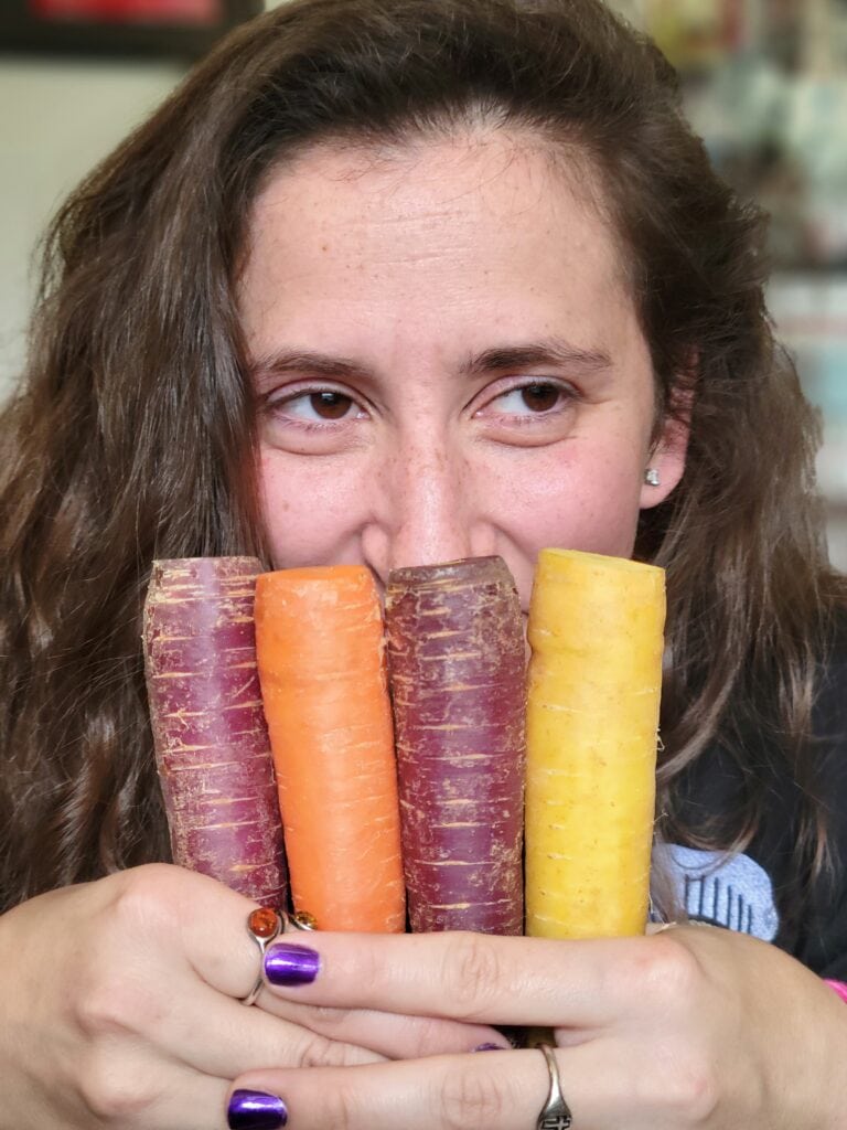 Woman holding purple, orange, and yellow carrots in front of her face while smiling at the camera. Bright and playful business photography from a chef and food content shoot near me.