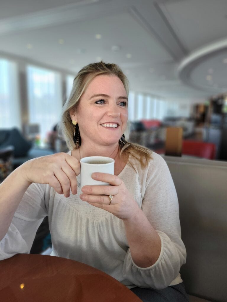 Blonde woman in a white blouse holding a white coffee mug while smiling and looking off camera in a bright lounge. Relaxed business photography image from a travel agent content shoot.