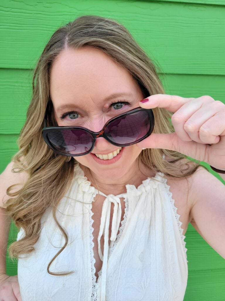 Woman in a white sleeveless top smiling and pulling down oversized sunglasses in front of a bright green wall. Playful business photography portrait captured during a travel agent content shoot.