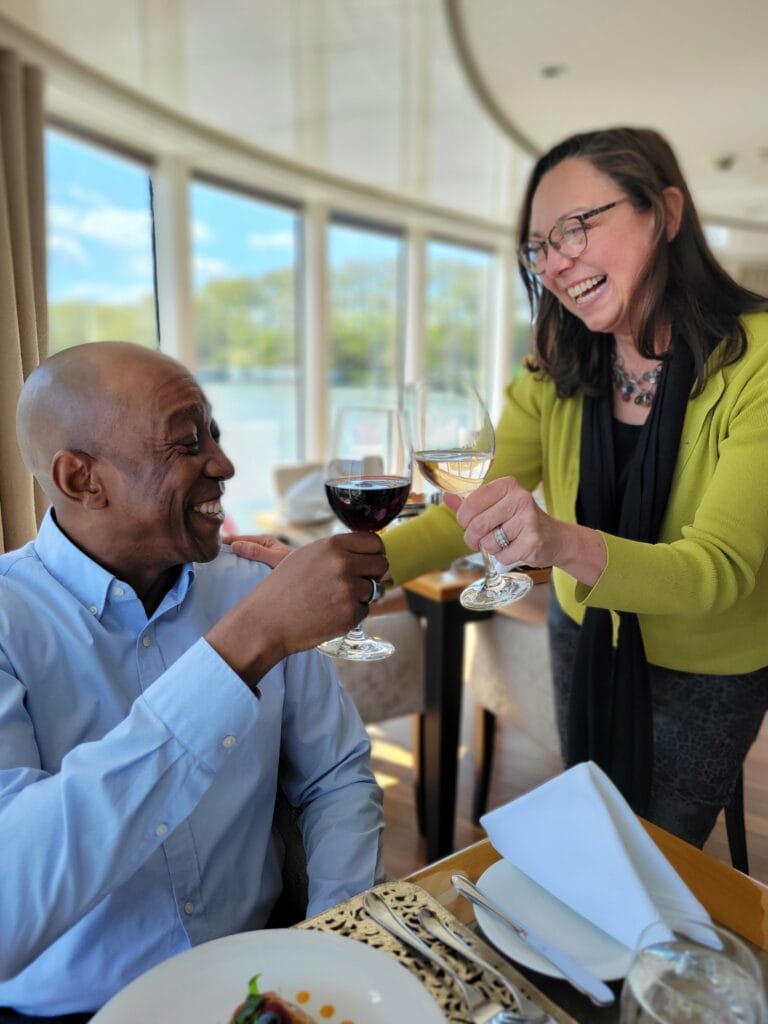 Two people smiling and toasting glasses of red and white wine at a restaurant table beside large windows. Elegant travel agent content shoot image showcasing luxury dining and connection.