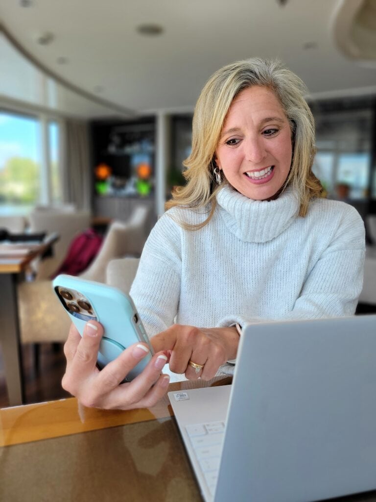Woman in a white turtleneck sweater smiling while looking at her phone beside an open laptop in a bright lounge. Modern business photography image for a travel agent brand photoshoot focused on digital communication.