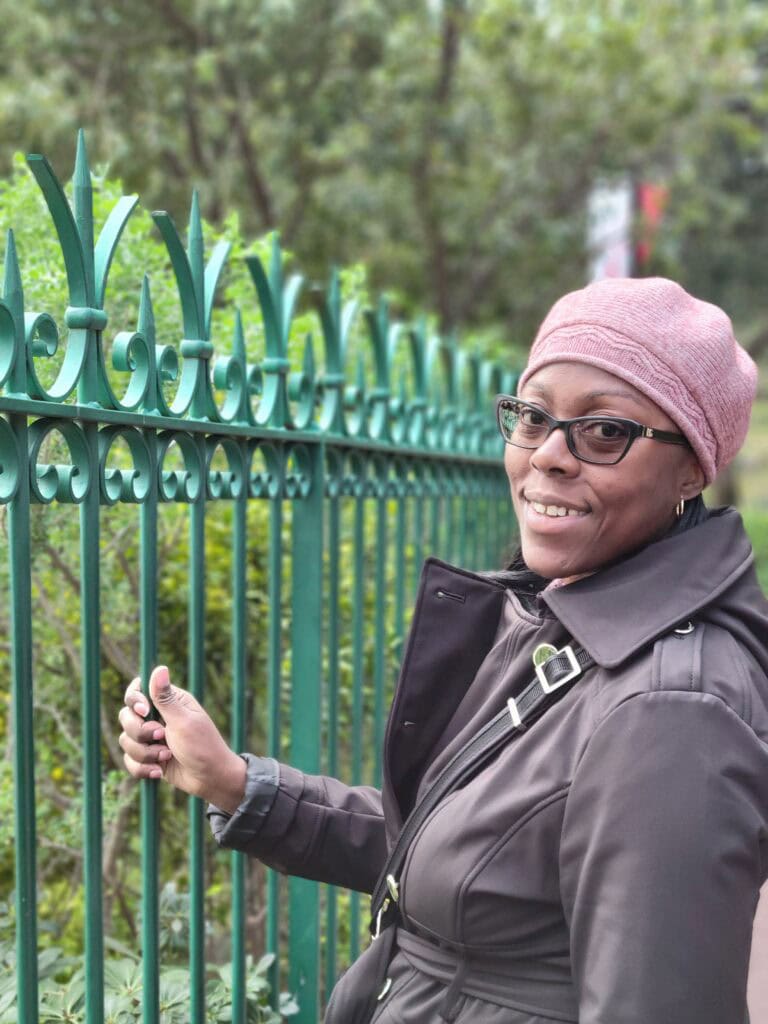Woman in a pink knit hat and dark coat smiling beside an ornate green metal fence surrounded by greenery. Travel agent content shoot portrait with a natural outdoor setting.