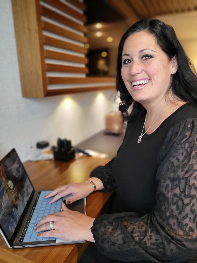 Woman in a black blouse with sheer sleeves smiling while typing on a laptop at a wooden desk in a modern lounge. Polished brand photoshoot image for travel agent content and business photography.