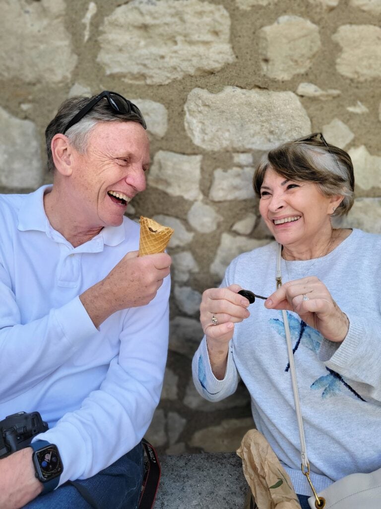 Senior couple sitting together outdoors and laughing while sharing ice cream and snacks in front of a stone wall. Lifestyle business photography image captured during a travel agent content shoot.