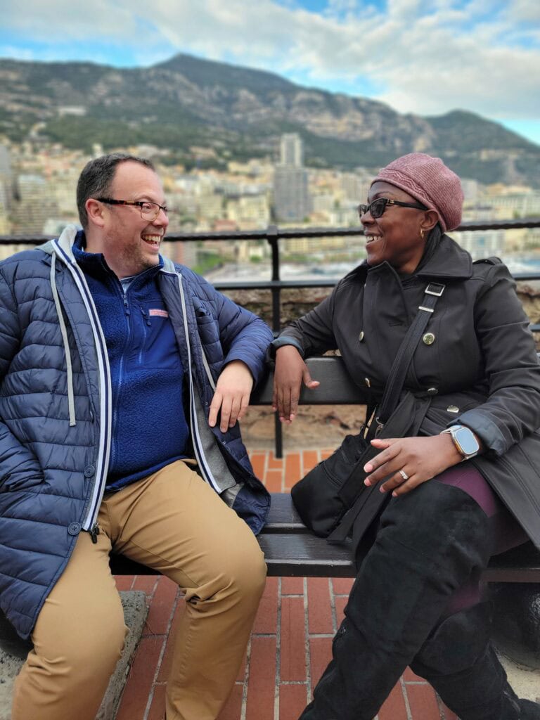 Two people in winter coats sitting on a bench and laughing together with mountains and a coastal city in the background. Lifestyle business photography captured during a travel agent content shoot abroad.