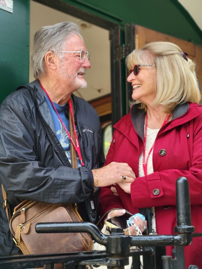 Older couple in jackets smiling and holding hands while standing together outside near a train car entrance. Travel agent content shoot image highlighting connection and shared experiences during travel.
