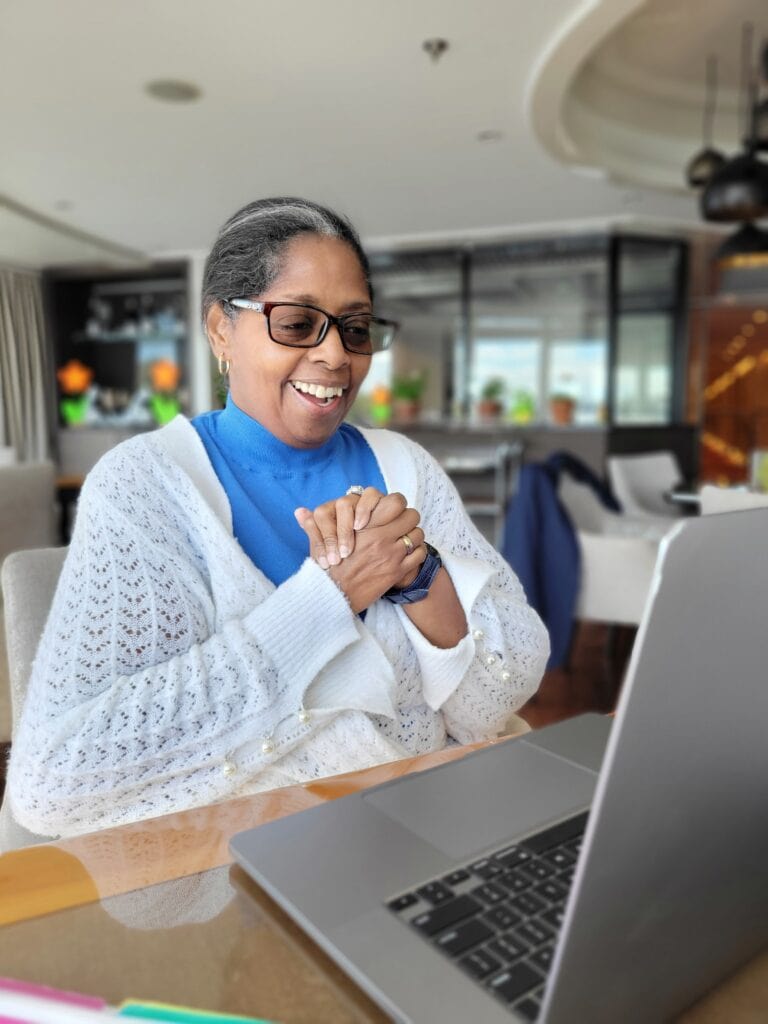 Woman in a white cardigan and blue top smiling at a laptop during an online meeting at a polished workspace. Professional business photography for a travel agent content shoot and brand photoshoot.