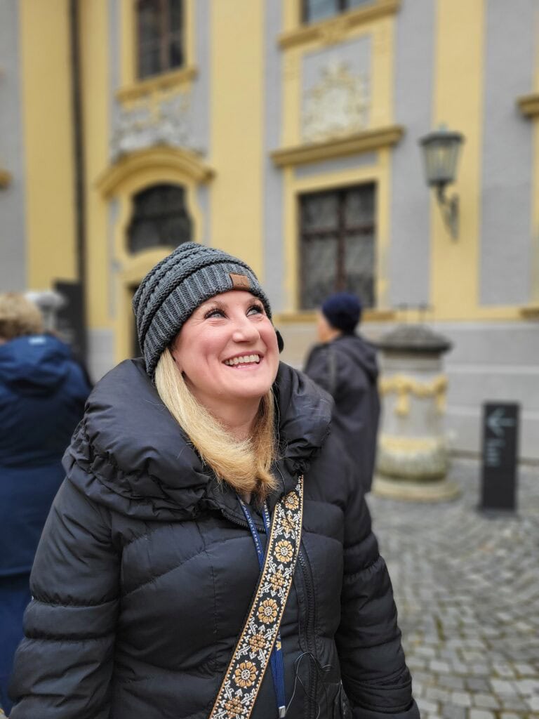 Smiling woman in a black winter coat and gray knit hat standing on a cobblestone street in front of a historic European building. Travel agent content shoot photo highlighting cold weather travel experiences and destination storytelling.