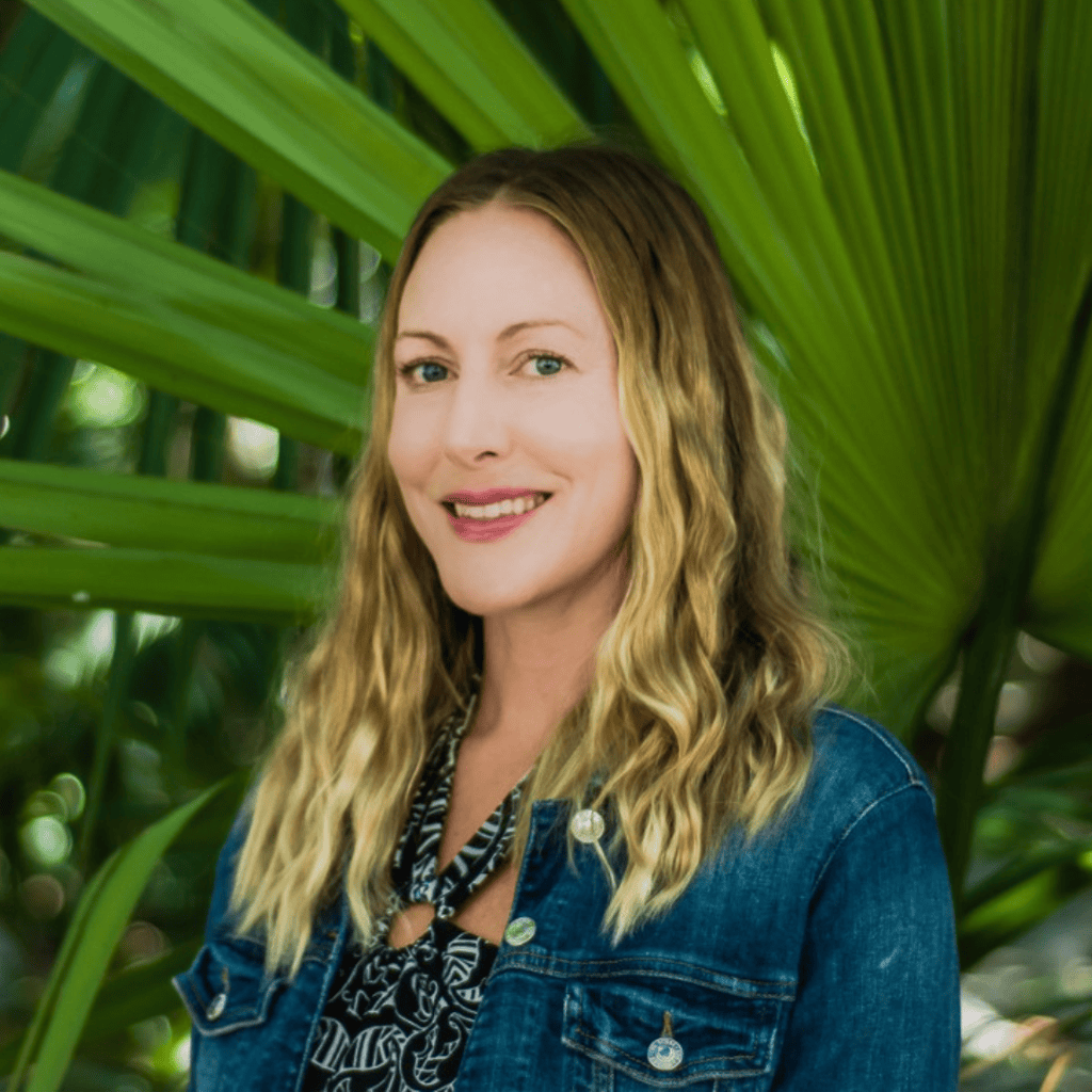 Jamie Rose smiles at the camera wearing a denim jacket with lush green tropical leaves in the background. Jamie is the Director of Business Development and Partnerships at Tembo Wild.